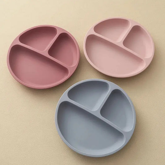 Three round three-compartment divided plates in dusty pink, light pink, and light blue-gray, arranged on a warm beige textured background.