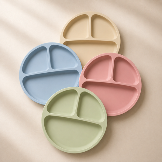 Four round divided baby plates in pastel shades of beige, blue, pink, and sage green, arranged in a fan-like composition on a warm beige background with soft diagonal shadows.