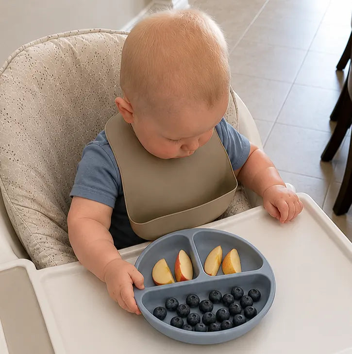 A baby sitting in a high chair wearing a beige bib and blue shirt, holding a gray divided plate with sliced apples and blueberries on a white tray.