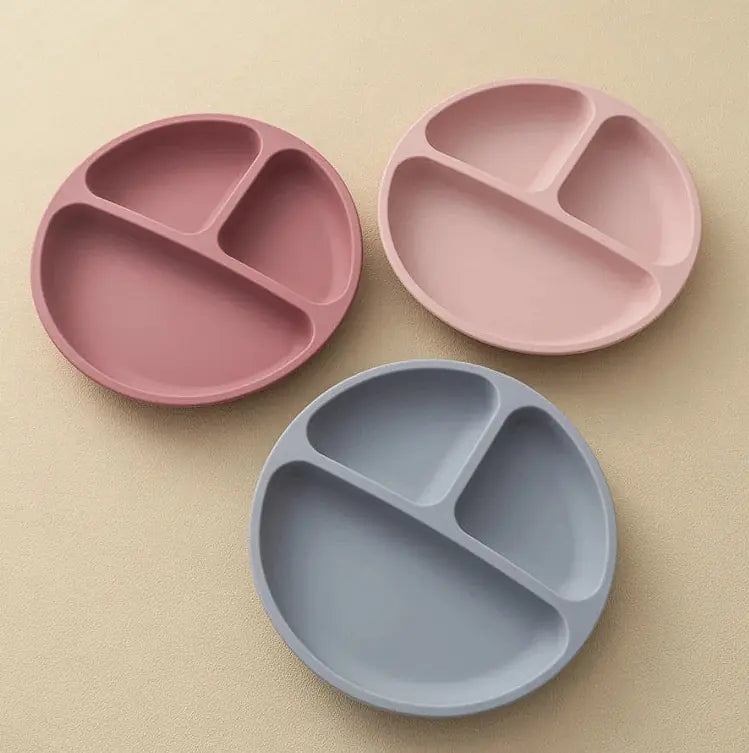 Three round three-compartment divided plates in dusty pink, light pink, and light blue-gray, arranged on a warm beige textured background.