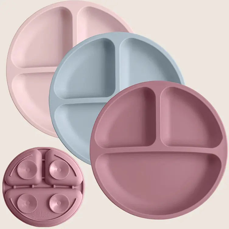 Four silicone divided baby plates in soft pastel colors, including a large pink plate, a large dusty rose plate, a large light blue plate, and a smaller matching pink plate shown folded flat with suction cups, on a light beige background.